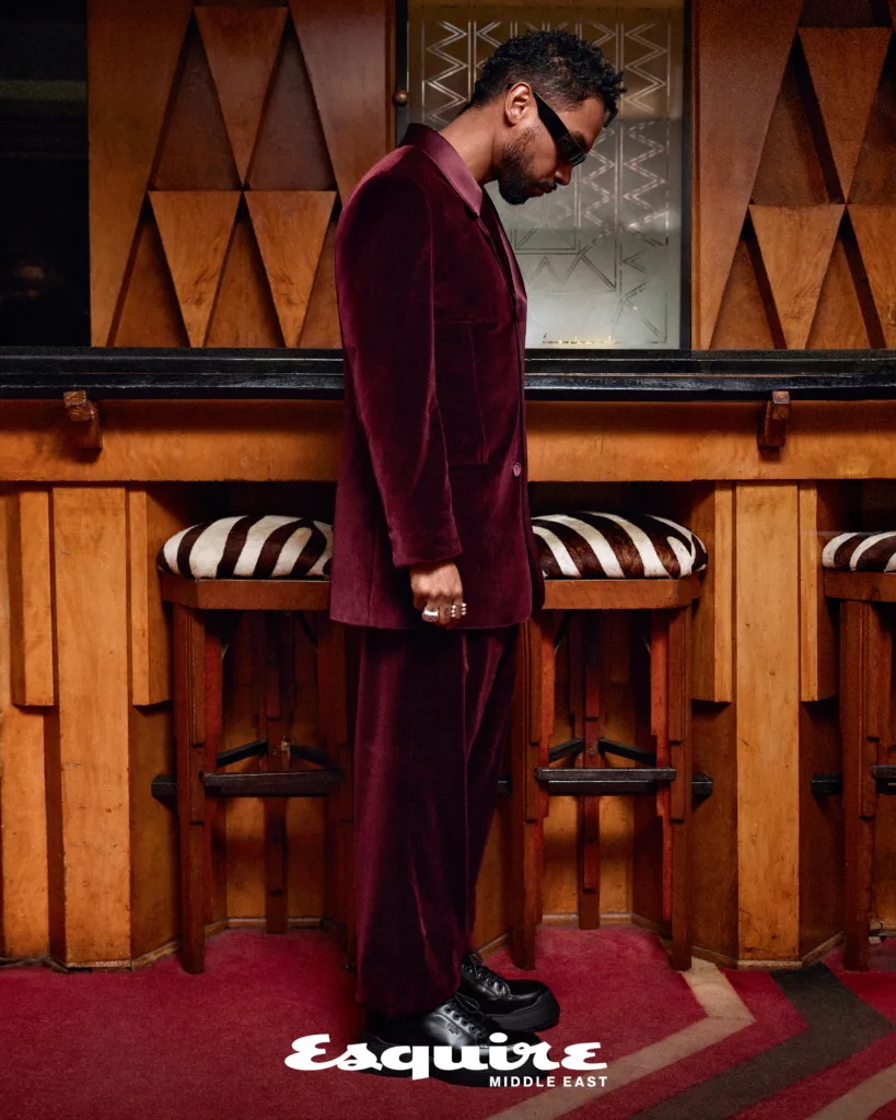 US singer Miguel standing side profile in a burgundy Fendi suit in a room with wooden art deco furniture