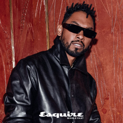 US singer Miguel wearing black leather Fendi jacket and pants and black sunglasses leaning against a red wall while smiling
